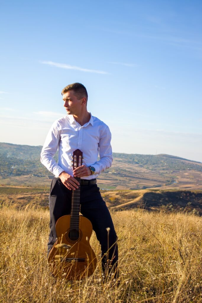 Home Young man in white shirt holding guitar in grassy field under clear blue sky.